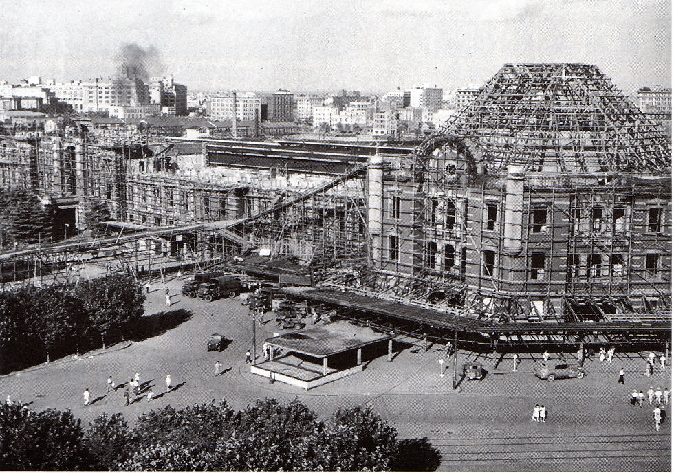 Large building under construction with scaffolding, surrounded by people and vehicles. Cityscape in background. Black and white.