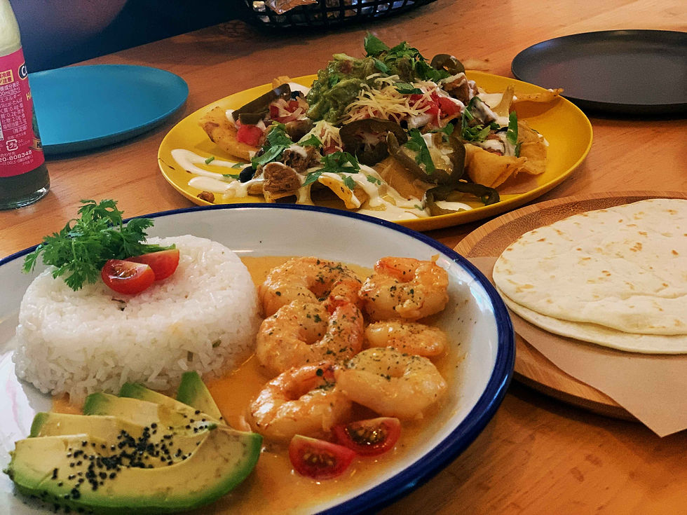 Shrimp with rice, avocado, and salad on plates. Colorful nachos and tortillas nearby on a wooden table. Casual dining setting.