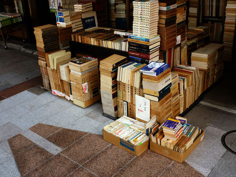 Stacks of books and boxes are displayed on the pavement in a dimly lit space with visible price tags, creating a calm and curious mood.
