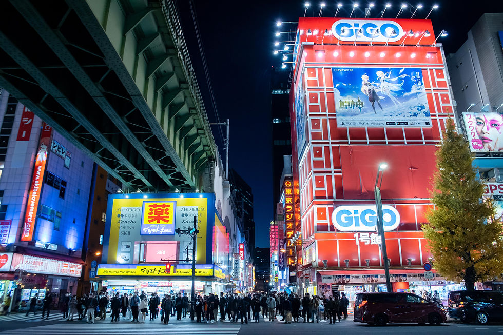 Busy nighttime city street with a crowd crossing. Brightly lit red and yellow storefronts, including "GiGO," under an overpass.