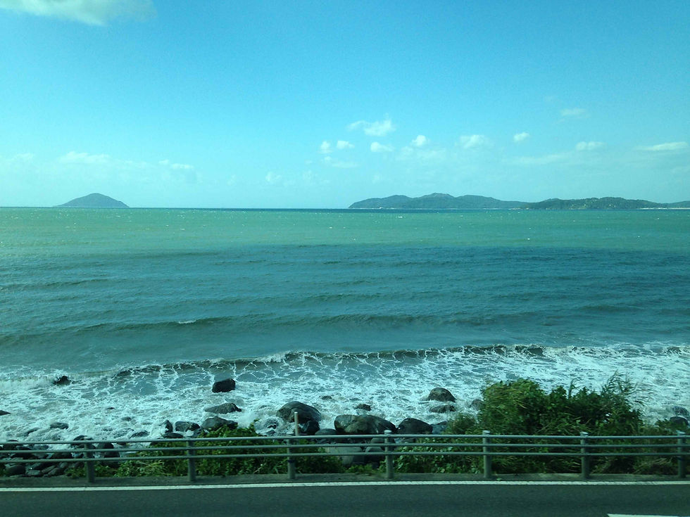 Ocean waves crash on rocks by a roadside. Distant green islands under a blue sky evoke a calm and serene mood.