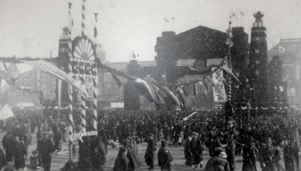 A large crowd gathers in a city square adorned with flags and banners. The setting is busy and festive, with a historical ambiance.