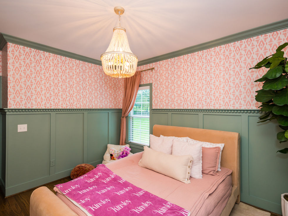Cozy bedroom with a pink-patterned wall, chandelier, green panels, and plants. Bed has plush pillows and a blanket with "Kinsley" text.