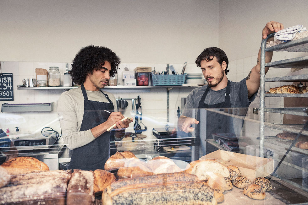 Two bakers in aprons at a bakery counter. One takes notes while the other arranges bread on racks. Kitchen tools in the background.