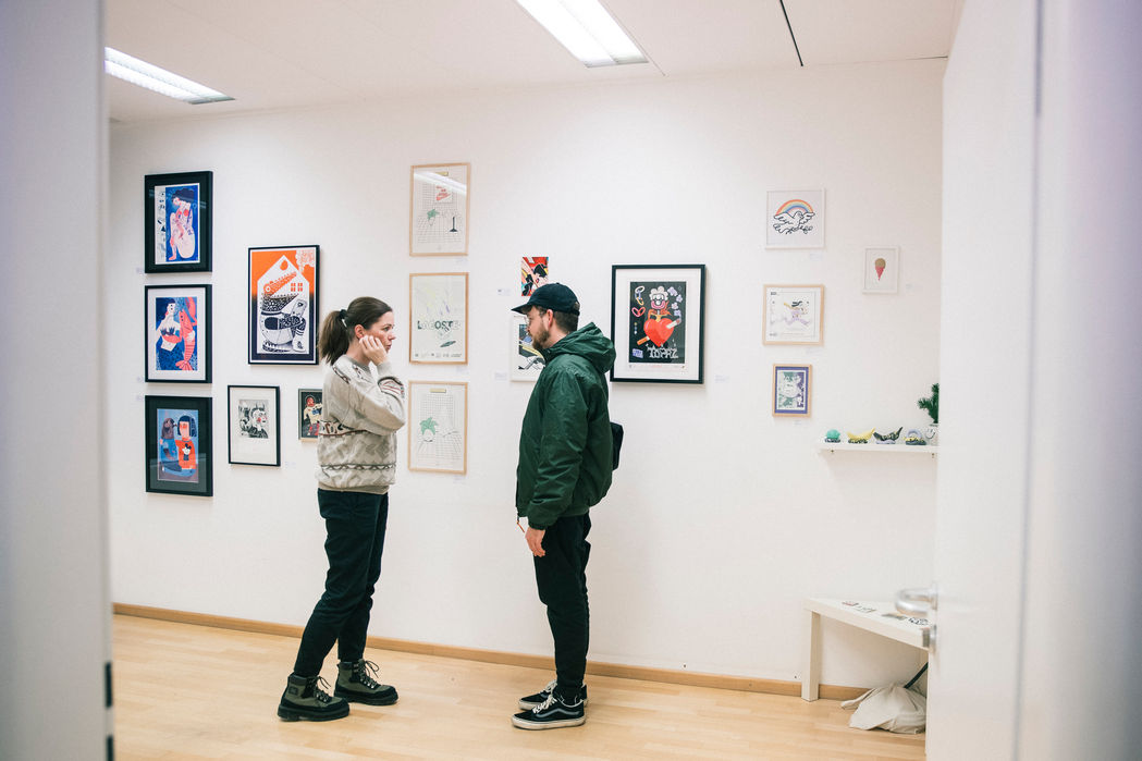 Kunstausstellung: Zwei Personen betrachten eine Wand voller gerahmter, moderner Illustrationen.