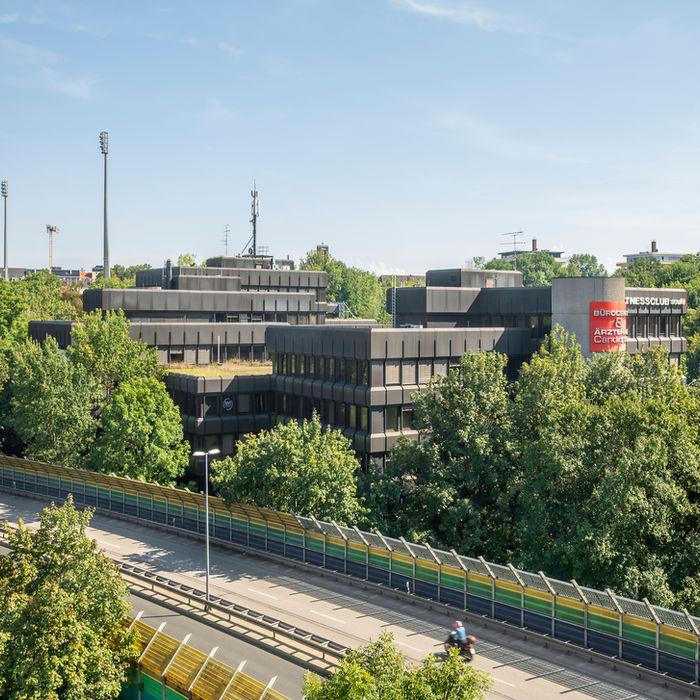 Panoramaaufnahme eines Bürogebäudes umgeben von viel Grün und einer Autobahn im Vordergrund.