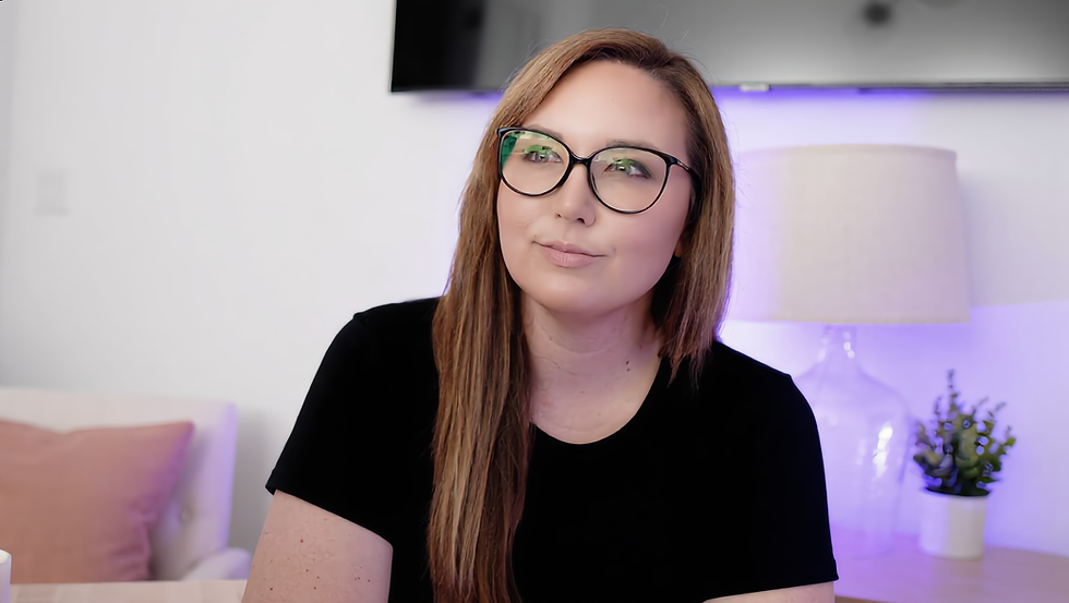Headshot of a woman with long auburn hair and black-framed glasses, wearing a black t-shirt, with a softly lit purple background behind her.
