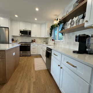 A modern kitchen featuring sleek white cabinetry, stainless steel appliances, and quartz countertops with a subtle marble pattern. The spacious island offers barstool seating, while under-cabinet LED lighting and natural light from large windows enhance the open, airy atmosphere.