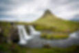 Iconic Kirkjufell mountain in Iceland with cascading waterfalls in the foreground, showcasing the country's dramatic natural landscape. The green mountain rises distinctively against cloudy skies, with flowing water representing Iceland's renewable energy resources.