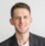 Headshot picture of Jacob Norris. Smiling man with short wavu hair in a black blazer and white shirt. Neutral gray background, conveying a professional, friendly mood.
