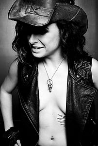 Mistress Wolfe in a leather vest and leather cowboy hat, shirtless underneath. Eyes downcast with a slight grin, oozing Daddy energy. Shot by Mark Dektor