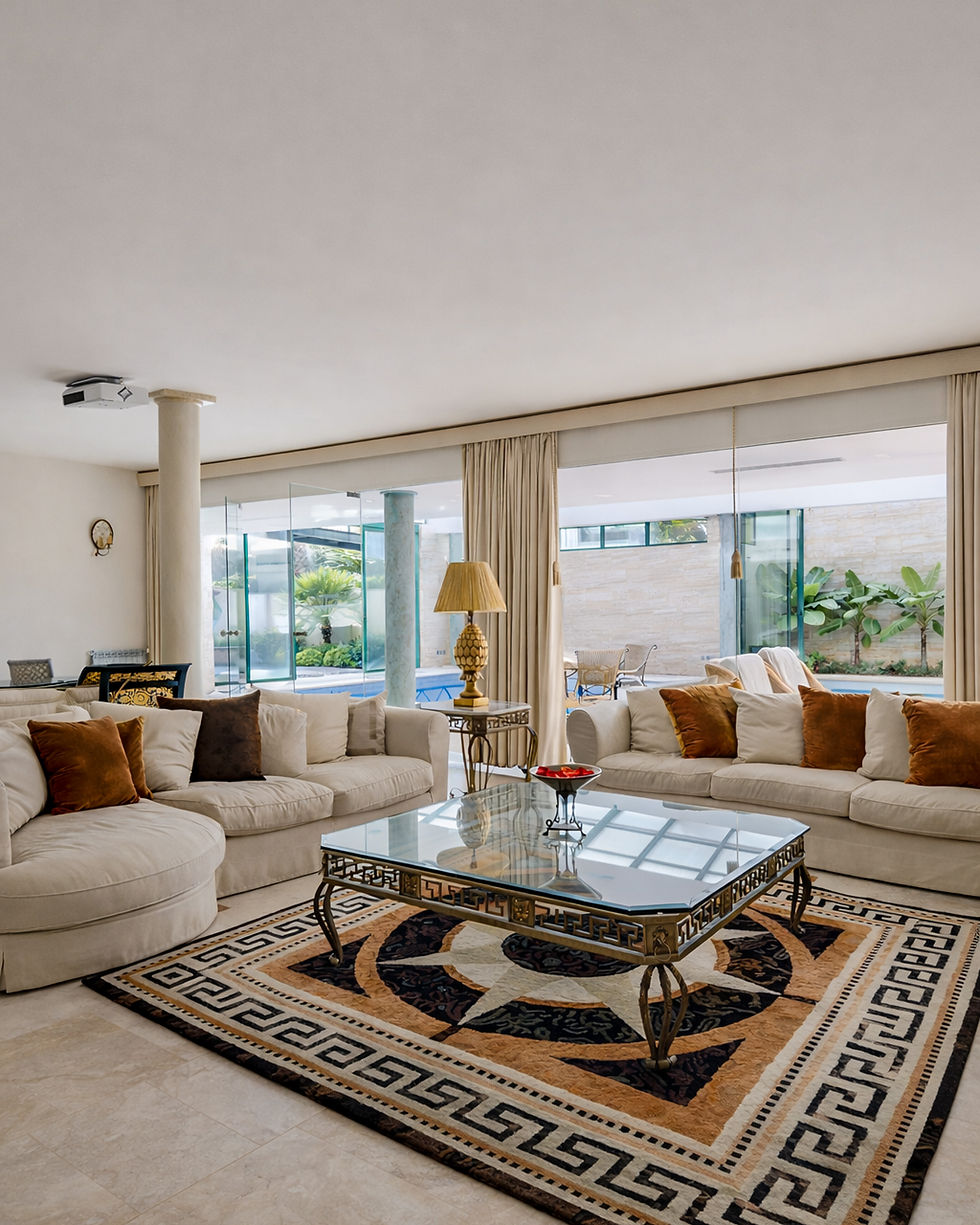 Spacious living room in a private residence in Alvalade, Lisbon, featuring neutral-toned sofas, a glass-topped coffee table with classical detailing, patterned area rug, soft ambient lighting, and floor-to-ceiling glass doors opening onto a landscaped indoor patio.