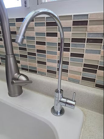 Grifo cromado en cocina con azulejos de colores neutros de fondo. Mostrador claro con fregadero blanco a la izquierda. Ambiente moderno. Grifo de agua Rainsoft instalado en la cocina de una vivienda en Puerto Rico