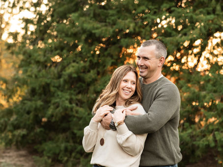 strawberry blonde wife stands infront of her husband who has wrapped his arms around her shoulders. She is in a cream sweater and cuddling back into his chest. They both have large smiles with teeth showing. There are fall foliage trees behind them.