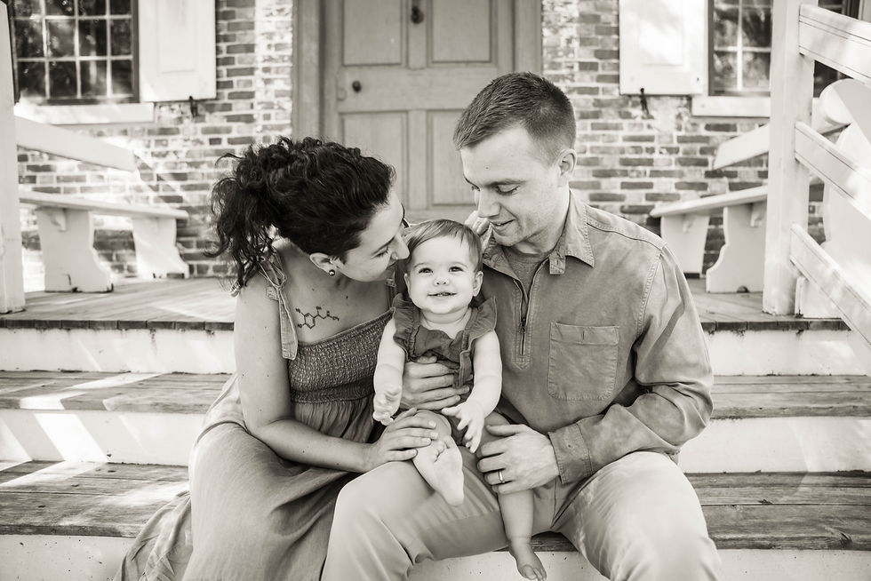 Black and white photograph of parents sitting on steps with the baby on dads lap between them. Mom is smooching her daughters hair and baby has the biggest smile.