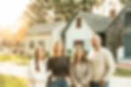 the family of four is standing in front of the historic buildings in the society's park with three buildings in the background. The youngest daughter starts the line of hte family on the far left and her arms are wrapped around her moms arm. Next to the mom is the oldest daughter who is shorter than the mom and dad. Then the dad is on the far right of the family line.