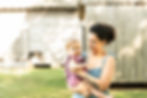 Mom is holding her baby on her hip. Baby is looking towards the camera and mom is looking down at baby. There is a wooden building in the distance behind them. The little girl has a smirk of happiness on her face.