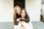 Mom is standing behind her tween daughter with her arms wrapped around her daughters shoulders. The daughter has lifted her arms to embrace her mother hugging on her. Both are smiling with big natural smiles.