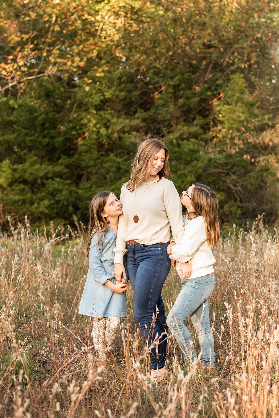 Mom is standing in between her two daughters while holding their hands. Mom is standing taller than both girls by 14 inches. Mom is looking down at her daughter to her right side. Another daughter is standing on moms left and is looking up at her. There is green and yellow foliage behind them in the distance. 