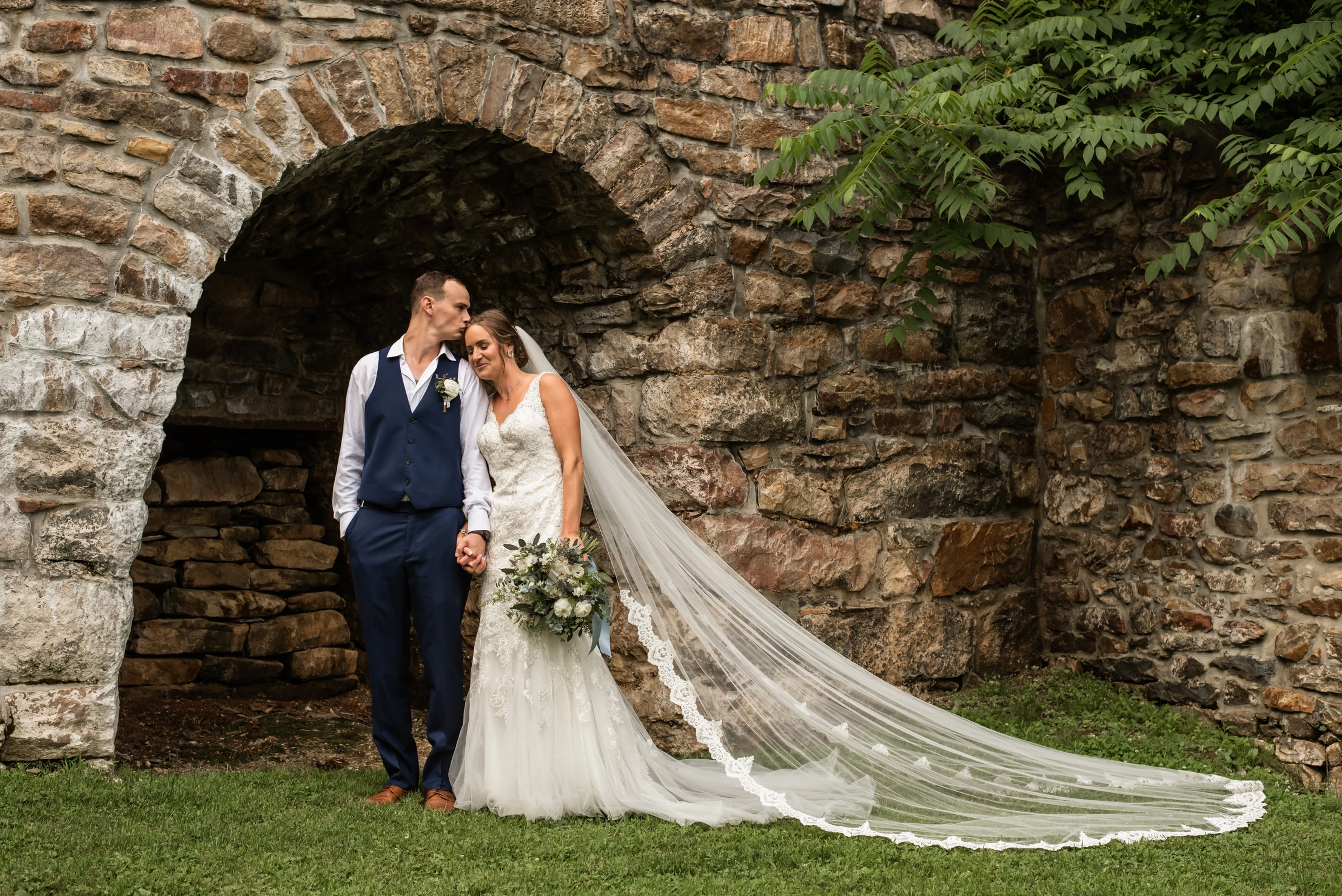 Bally Spring Inn bride is leaning on her husbands shoulder while he kisses her forehead. The brides long cathedral length viel is flowing behind her. There is a large historic stone wall behind the couple and they are centered in the arch.