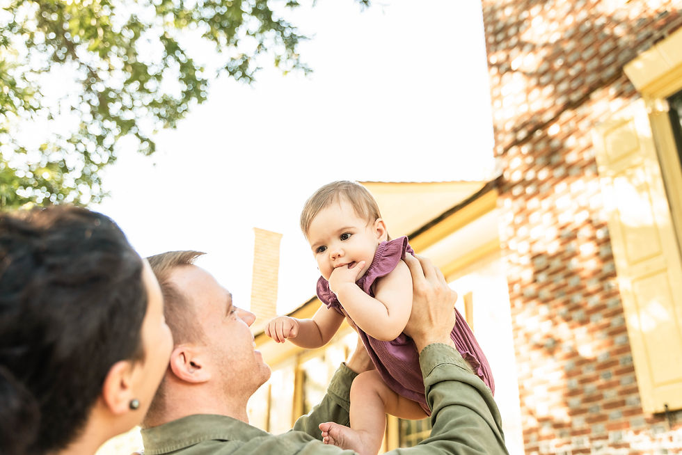 Mom cuddling on dads back while dad lifts little one above his head. Baby has her fingers in her mouth.