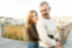 Wife is standing on the Lewes boat yard dock behind her husband. Her arms are wrapped around his waist. The husband is looking back to her. Both of them are smiling with their teeth showing.