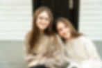 The historic lewes porch in the historic society has two sisters sitting next to each other with a black door behind them. The younger sister is leaning on the shoulder of her older sister.
