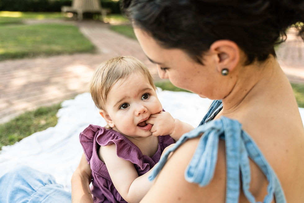 Mom is sitting on white blanket in the middle of a brick circle. The camera is looking down over moms shoulder to her daughter. The daughter is looking up towards mom which shows her ruffled sleeve and mom's profile looking at her.