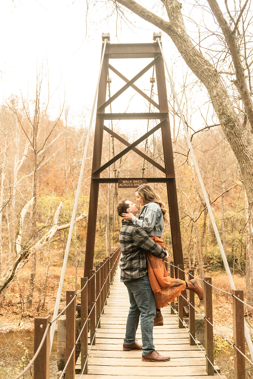 This 2025 wedding couple are embracing on a wooden bridge in autumn forest during their engagement session with Shannon Ritter Photography, a Delaware based (not bound) wedding photographer. Billy is holding onto his fiance surrounded by warm colors, and a romantic mood.