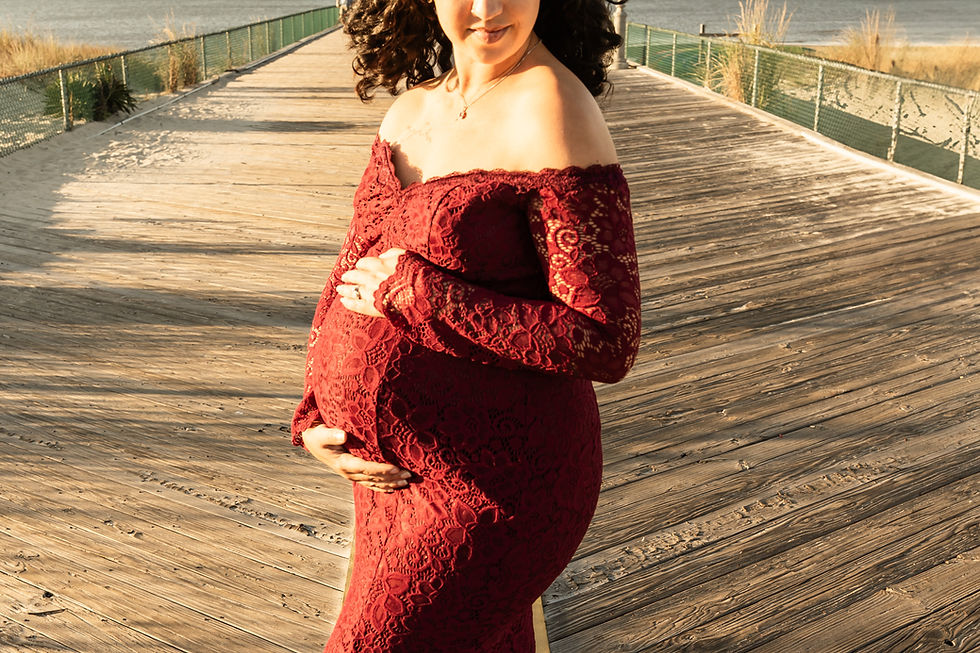 Pregnant woman in red lace dress on Cape Henlopen fishing pier cradles belly on a wooden boardwalk by the sea, under warm sunlight, expressing tranquility.