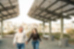 Real moment of husband and wife holding hands between them running towards the camera while the couples looks towards each other giggling. Over their heads you see the pergola style open roofing framing them.