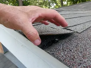 A hand lifting a loose asphalt shingle on an eave to check the home's roof integrity during a routine inspection.