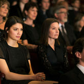 Audience in formal attire sits attentively in a dimly lit theater. Focused expressions suggest anticipation or reflection.