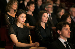 Audience in formal attire sits attentively in a dimly lit theater. Focused expressions suggest anticipation or reflection.