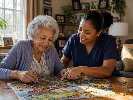 An elderly woman with silver curly hair and a light cardigan smiles while working on a jigsaw puzzle with a warm, engaged female caregiver beside her in a sunlit, cozy living room.
