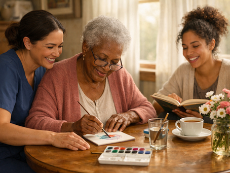 Three smiling women paint and read together at a sunlit table, with tea, flowers, and a watercolor set in a cozy home.