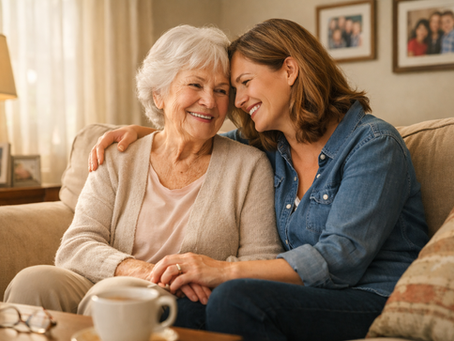 Two women, one elderly and one middle-aged, sit on a cozy couch, smiling and embracing. Warm lighting, with family photos in the background.