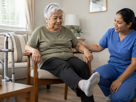 Elderly stroke survivor with short gray hair performs a seated leg exercise at home while a female caregiver in plain blue scrubs provides gentle support in a bright living room.