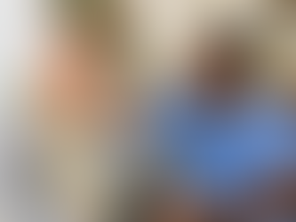 Elderly man drinks water while sitting on a sofa. A caregiver in a blue uniform smiles beside him.