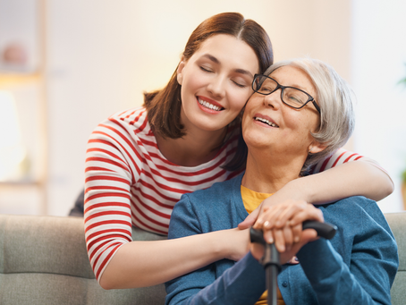 A daughter is affectionately embraces her mother from behind as they both smile warmly while sitting on a couch.
