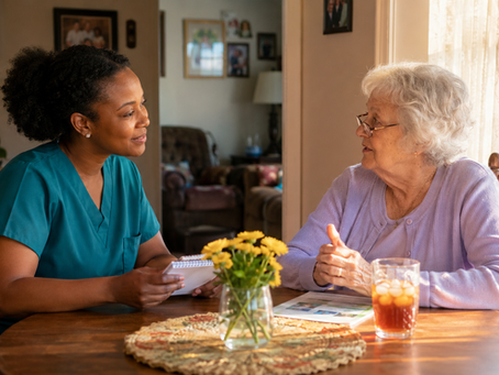 A Black female caregiver in teal scrubs with natural hair pulled back listens attentively and takes notes at a sunlit kitchen table across from an elderly white woman with short silver hair and a lavender cardigan as they discuss her personalized home care plan together.