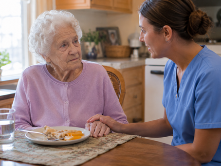 A caregiver in blue scrubs sits beside an elderly woman in a lavender cardigan at a sunlit kitchen table, gently holding her hand while she eats a meal of soft, cut-up food.