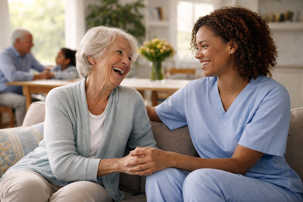 Elderly woman and caregiver in blue scrubs smile joyfully on a couch. Background shows a man and child at a table, plants, and flowers.