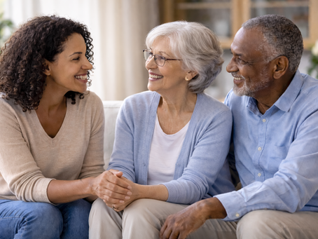Three people sit smiling; a younger woman holding hands with an older woman, and an older man. They sit on a couch in a bright room.
