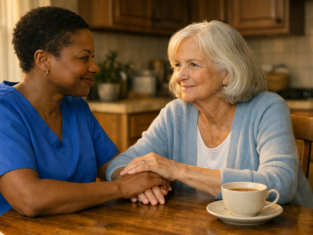 A middle-aged Black female caregiver with short natural hair and a burgundy blouse sits at a sunlit wooden kitchen table, her hand rested upon by an elderly white woman with silver hair and a pale blue cardigan, sharing a quiet and tender moment of mutual connection.