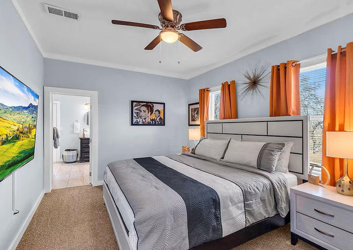 Bright bedroom with king sized bed, TV, two windows, and end tables.  The color scheme is white, black, and orange.