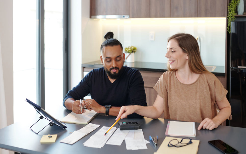 A woman and man sit at a table with papers and a calculator, appearing focused and cooperative. Bright kitchen background.