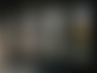 Sunlight streams through four tall windows onto a dark, empty room with a shadowed floor. Outside, buildings and trees are visible.