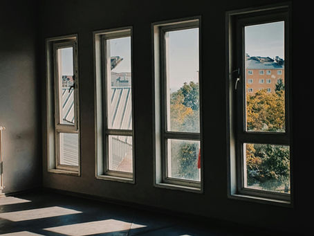 Sunlight streams through four tall windows onto a dark, empty room with a shadowed floor. Outside, buildings and trees are visible.