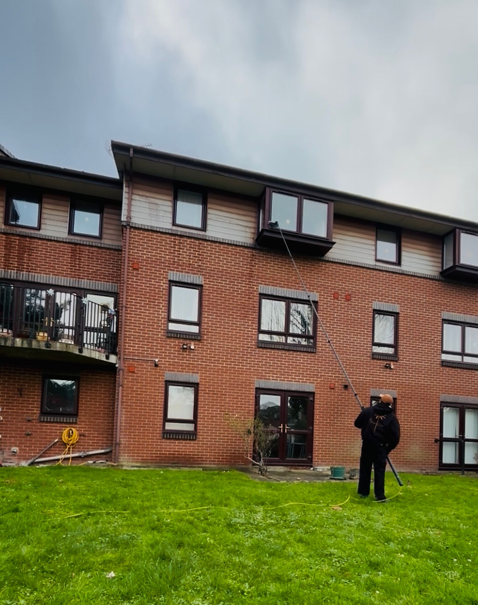 A window cleaning professional from The Riviera Group stands on a grassy lawn below a brick apartment block, holding a long pole to clean the exterior windows of a building.